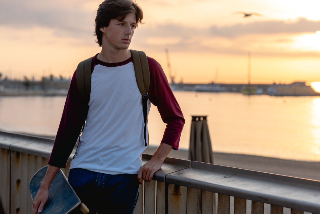 A boy wearing a raglan t-shirt stands by a river at sunset