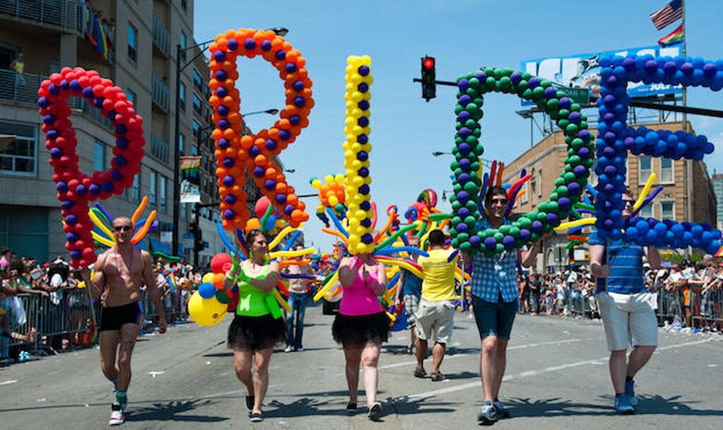People marching while carrying balloon structures which spell "pride"
