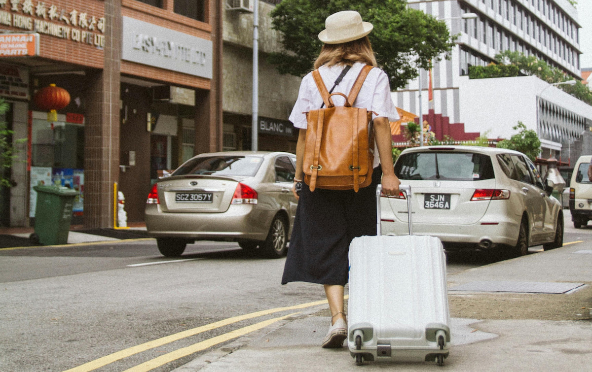 A woman walks in a city with a roll-along suitcase and a small backpack
