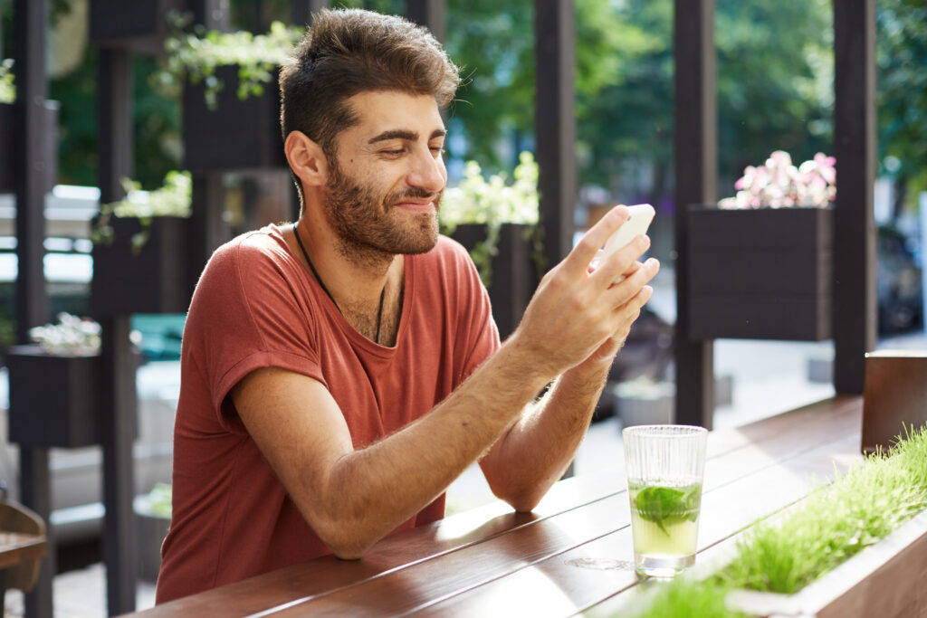 A smiling man sits on a bench outside while looking at his phone